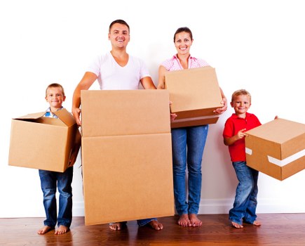 A family of four, composed of two adults and two children, is standing indoors on a wooden floor in front of a plain white wall. The adults are positioned centrally, holding large cardboard boxes; the man on the left is wearing a white t-shirt and jeans, while the woman on the right is dressed in a pink checkered shirt and jeans. The children, a girl and a boy, are standing on either side of the adults, each holding smaller cardboard boxes. The boy on the far right is wearing a red t-shirt and jeans, holding a medium-sized cardboard box, while the girl on the far left is wearing a blue t-shirt and jeans, holding a slightly larger box. The scene is well-lit, indicating a bright, natural indoor environment, typical of a residential setting in Notting Hill during a house move. The family appears ready to load or unload moving boxes, with a focus on household belongings, which suggests they are engaged in a local removals or relocation activity, supported by the professional house removals service of Notting Hill Man with Van.