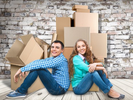 A young man and woman sit back-to-back on a light wooden floor in front of a whitewashed exposed brick wall, surrounded by multiple cardboard moving boxes of various sizes, some sealed and others open with flaps partially lifted. The man has short dark hair and is dressed in a blue and white checkered shirt with dark jeans and sneakers, smiling and leaning slightly to his left. The woman, with long wavy red hair, wears a turquoise sweater over a collared shirt and blue jeans, also smiling and with her arms crossed. Behind them, the boxes are irregularly stacked, with some leaning against each other, indicating a recent move within Notting Hill or nearby London districts. The scene suggests an active house or flat move, with the furniture and boxes arranged on a spacious interior floor, likely part of a residential property in a typical London neighborhood, as referenced by the brick wall and the urban setting. The lighting is bright and natural, emphasizing the details of the cardboard textures and the cheerful expressions of the individuals, fitting a professional removals service context.