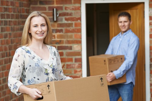 A young woman with long blonde hair, wearing a light-colored floral blouse, smiling while holding a medium-sized cardboard box with 'UP' arrows on the side, standing outside a residential property with a brick wall and a wooden door frame visible behind her. In the background, a young man with short brown hair, dressed in a blue button-up shirt and jeans, is standing on the doorstep, holding a larger cardboard box. The scene is set on a residential street in Notting Hill, with natural daylight illuminating the scene, suggesting a professional house removal or relocation activity by Notting Hill Man with Van. The interior of the house can be partially seen through the open door, indicating the context of moving household items during a house removal process.