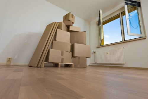 A newly moved into empty room with light wooden flooring and white walls, showing a stack of cardboard boxes of various sizes placed on a wooden pallet in the corner near a radiator. To the left of the boxes, there are several flat-packed items, possibly furniture components, leaning against the wall. The room has a large, open window on the right side, with a view of a clear blue sky and the outdoors. The window is open, allowing natural light to illuminate the space, highlighting the clean and minimal environment. The scene depicts the early stages of a house move managed by Notting Hill Man with Van, with household items temporarily stored in the room ahead of further furniture and personal belongings transportation. The overall setting suggests a residential property in a quiet area, typical of a house or apartment in Notting Hill or nearby in West London, with a focus on safety and careful handling during the removal process.