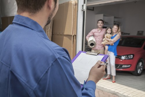 A professional removals worker, dressed in a blue shirt, is seen from behind holding a clipboard and taking notes or checking a checklist outdoors in a residential driveway. In the background, a family of three — a man, woman, and young child — are standing near the entrance of a house, smiling and looking toward the camera. The man is wearing a pink shirt, the woman is in a blue dress, and the child is dressed in beige. To the right, a red car is parked close to the garage door, which is partly open, revealing some household items inside. The driveway is paved, and the setting suggests a typical UK suburban property, with elements indicating a house removal or relocation activity. The scene captures a moment of organization and preparation for a house move involving Notting Hill Man with Van, a local removals service provider noted in the context. The natural lighting indicates daytime, contributing to a clear view of all elements in the scene.