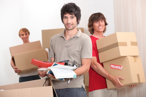 A group of three young men are indoors, preparing for a house move, with a plain white wall in the background. The man in the foreground has dark, slightly curly hair and a trimmed beard, wearing a light grey polo shirt, and is holding a few notebooks, a clipboard, and a black pen, looking directly at the camera. To his right, a man with medium-length brown hair and a red t-shirt is smiling and carrying two medium-sized cardboard boxes, one labeled 'Fragile' with brown packing tape. In the background, a third man with short blond hair, wearing an off-white shirt, is holding a single cardboard box with both hands. The room appears well-lit with natural light, and the scene reflects typical household packing activities, with assorted cardboard boxes, some sealed with tape, ready for relocation, consistent with house removal services in Notting Hill or the surrounding London area.