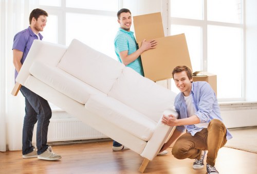 Three men are engaged in a house removal activity inside a bright, modern room with large windows and white walls. The man on the right, wearing a light blue shirt and brown trousers, is crouched down and holding the armrest of a white, fabric-upholstered sofa, which is being carefully moved. Behind him, another man in a turquoise striped shirt is carrying a medium-sized cardboard box, positioned slightly to the right of the scene. A third man, dressed in a purple shirt and dark trousers, stands to the left, assisting or observing the moving process. The sofa appears to be partially lifted or positioned for transport, with the length extending diagonally across the room. The room has a minimalist look with a clean wooden floor, and the large windows in the background suggest a bright, airy environment, typical of a residential property in Notting Hill or nearby west London. This scene visually captures the effort involved in house removals, focusing on teamwork, furniture handling, and the use of boxes during a relocation process with a professional removals company, Notting Hill Man with Van.