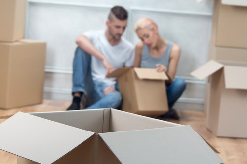 A man and woman are seated on a light wooden floor inside a residential space, surrounded by numerous cardboard moving boxes in various sizes, some open and some closed, with a few stacked against the wall. The man has short dark hair and a beard, wearing a white T-shirt and blue jeans, and is holding an open box, while the woman, with blonde hair tied back and dressed in a grey sleeveless top, is looking inside another box. The background shows a plain grey wall and a row of stacked cardboard boxes, indicating an organized packing process typical of household removals in a Notting Hill area residence. The lighting is natural, suggesting daytime, and the setting appears to be a living room or similar part of a residential property preparing for moving. The scene emphasizes careful packing and handling of household items, consistent with professional removals services offered by Notting Hill Man with Van.