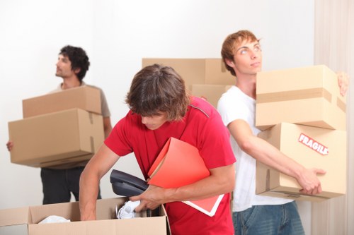 Three movers from Notting Hill Man with Van are in a residential house interior preparing for a move in Notting Hill. The mover in the foreground, wearing a red t-shirt, is leaning into an open cardboard box with packing materials and a black handheld device, holding a bright red folder. In the background, a man with curly dark hair, dressed in a light grey t-shirt, is carrying a medium-sized cardboard box labeled 'fragile,' with his back turned towards the camera. To the right, another mover with blond hair, wearing a white t-shirt, is holding a cardboard box in each arm, positioned near a doorway or corridor within a house. The scene depicts a typical house relocation setup with multiple boxes of various sizes stacked or being carried, within a clean, well-lit interior. The image reflects professional moving services, subtly hinting at local household removals in the Notting Hill area, with a focus on careful handling of personal belongings and efficient packing. The environment appears organized, with cardboard boxes, some sealed with packing tape, and individuals actively engaged in the moving process.