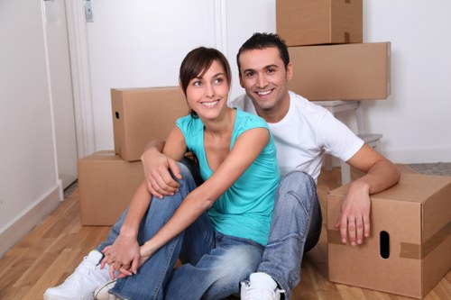 A smiling couple sits on a light wooden floor inside a residential property, surrounded by several cardboard moving boxes of various sizes, some stacked in the background near a white wall and a section of a staircase. The woman, wearing a bright turquoise top and jeans, is seated cross-legged, leaning slightly on the man, who is dressed in a white T-shirt and light blue jeans. The scene appears to depict a house relocation, with the couple preparing for or in the process of moving, as suggested by the unpacked boxes and casual posture. The environment is well-lit with natural or indoor lighting, and the setting suggests a typical household interior in London, close to Notting Hill, consistent with the local SEO context for a house removals service. Notting Hill Man with Van offers professional removals, and this image captures a moment of transition, emphasizing the preparation and organization involved in moving house within the area.