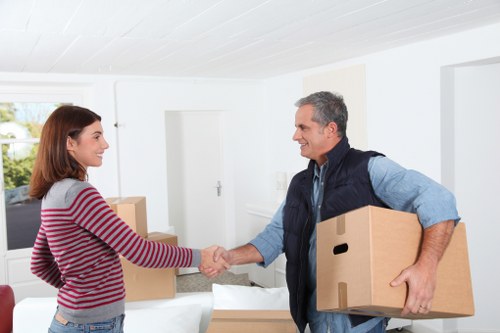 In a bright residential interior with white walls and a large window letting in natural light, a male mover from Notting Hill Man with Van, dressed in a blue shirt and navy vest, is smiling as he shakes hands with a woman in a striped red and grey sweater. The mover is holding a medium-sized cardboard box in his left hand and another box tucked under his right arm. In the background, there are additional cardboard boxes on a white surface, suggesting a moving or relocation process within a house in Notting Hill. The scene captures a professional and cordial moment during a house removal, highlighting the company's house removals service, with furniture and packing items possibly being prepared for transport. The environment appears clean and organized, emphasizing a smooth transition typical of house moving services in central London neighborhoods like Notting Hill, with the focus on respectful customer interaction and careful handling of household belongings.