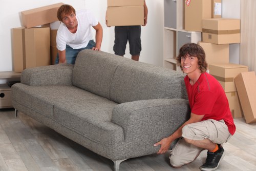 Two individuals are engaged in a house removal in a residential interior, possibly in Notting Hill or surrounding postcode area, with various cardboard boxes and household items visible around them. One person, wearing a white T-shirt, is kneeling behind a grey fabric sofa, seemingly positioning or stabilizing it. The other person, dressed in a red shirt and beige shorts, is crouching beside the sofa, holding and possibly moving the armrest section. The sofa is a modern, three-seater with a textured fabric finish, positioned on a wooden floor. The background features a white bookcase or storage unit filled with open and closed cardboard boxes of different sizes, some stacked at various heights, indicating an ongoing move. The room is well-lit, likely with natural light, emphasizing the clean, tidy environment typical of house removals. This scene captures the practical aspects of furniture handling and packing, with a focus on careful movement and organization, reflective of professional removals services in the London area.