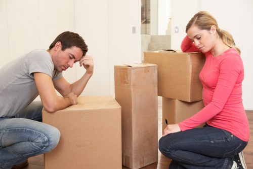 A man and a woman are seated on the floor of a residential interior, surrounded by stacked cardboard moving cartons and household items. The man, with dark hair, is wearing a light grey t-shirt and jeans, and appears to be resting his head on his hand while looking down at a box in front of him. The woman, with blonde hair, is dressed in a bright pink long-sleeved shirt and dark jeans, sitting on her heels with her head slightly tilted, looking contemplative. The scene is set in a well-lit, modern living room with neutral-colored walls and a staircase visible in the background, indicating they are preparing for a move within a Notting Hill or London residence. The presence of packed boxes suggests an ongoing house removal or relocation process, typical of a professional removals service such as Notting Hill Man with Van. The environment appears tidy and organized, with a focus on the packing stage of a home transition, emphasizing safety and careful handling of household belongings during the move.
