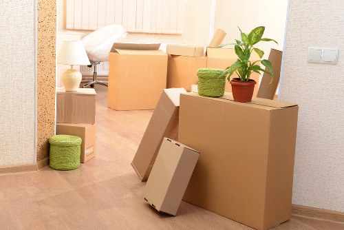 A scene inside a residential property with several cardboard moving boxes of various sizes, some sealed and others open, stacked or leaning against a wall in a hallway or living room area. The boxes are plain brown kraft paper, and some have visible flaps or packing tape. There is a small potted plant with green leaves on top of one of the larger boxes, adding a touch of greenery. To the left, a green woven storage basket is placed near the floor. In the background, a cream-colored sofa with cushions, a bedside table with a table lamp, and a wooden floor are visible, indicating a typical house interior in Notting Hill or nearby areas. The room is well-lit with natural or soft ambient lighting, and the overall scene suggests a house being prepared for moving or ongoing packing, consistent with professional removals services offered by Notting Hill Man with Van.