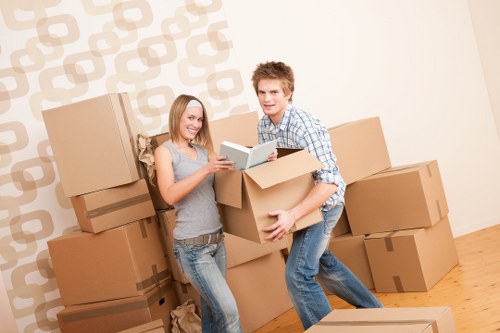 A young couple, a woman with shoulder-length blonde hair wearing a grey sleeveless top and jeans, and a man with short light brown hair wearing a checkered shirt and jeans, are standing indoors on a wooden floor surrounded by numerous brown cardboard moving boxes, some stacked against a wallpapered wall with a geometric beige and white pattern. They are smiling and appear to be preparing for a house move, with the woman holding a small open box and the man lifting a larger box, both engaged in packing or unpacking activities. The scene suggests an interior space such as a living room or hallway in a residential property, with natural lighting illuminating the scene. This image reflects typical household relocation activities carried out by a professional house removals company like Notting Hill Man with Van, serving areas including Notting Hill and surrounding parts of London, as part of their removals services focused on careful handling and efficient packing.