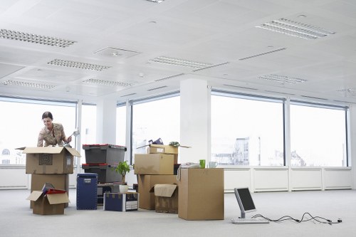 The image depicts a woman in a business suit packing or unpacking cardboard boxes in a bright, spacious office with large windows overlooking a cityscape. She is smiling and appears to be sealing a box with tape, with several other open boxes around her containing office supplies, paper, and small equipment. The boxes are of varying sizes, some stacked, and made of brown corrugated cardboard, with a few plastic storage containers and a small plant visible among them. The office interior features a plain white ceiling with recessed lighting, a radiator along the windowed wall, and a clean, minimalist environment. An office chair is placed on the floor near her, connected to a power outlet via an extension cord. The overall scene suggests active packing or moving preparations within a corporate office setting, consistent with house removals or office relocation services in London or nearby areas, which Notting Hill Man with Van might offer.