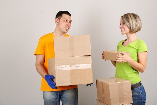 A young man wearing a bright yellow T-shirt and blue gloves is standing indoors in a residential setting, holding two cardboard boxes stacked vertically in front of him. He is looking towards a young woman, who is standing to his right, wearing a green T-shirt and jeans, smiling as she holds a smaller box with both hands. In the background, there are additional cardboard boxes placed on the floor, some sealed with packing tape, situated within a room that has plain, light-colored walls. The scene portrays a house removal or moving process taking place in a tidy, well-lit interior, possibly in a home in Notting Hill, London, with an emphasis on sustainable packing materials and careful handling of household belongings as part of environmentally conscious removal services by Notting Hill Man with Van.