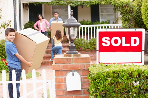 A family of four with light skin are engaged in a house move outside a residential property in Notting Hill, with a white picket fence and greenery surrounding the area. The father, wearing a light blue shirt, is carrying a large cardboard box towards the house, while the mother, dressed in a pink jacket, is walking behind him. A young girl with long hair, dressed in blue jeans and a grey top, is standing on the brick pathway near a classic lamp post, observing the activity. In the foreground, a young boy in a blue t-shirt is holding a medium-sized cardboard box with tape, positioned in front of a white-painted fence post. The house in the background has a white veranda with a railing, a front door, and windows with white shutters. To the right, there's a vivid red 'SOLD' sign mounted on a white background, partially obscured by lush green bushes, indicating the property has been recently sold. The scene is illuminated by natural daylight, capturing the moment of packing and organizing household belongings as part of a house removal process in a friendly suburban setting.
