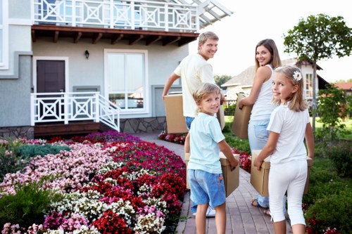 A family of four, consisting of a man, woman, and two young girls, are standing outdoors on a paved pathway in front of a residential house with a light grey exterior and white balcony railing. They are smiling and appear to be engaged in a house removal process, each holding cardboard boxes filled with belongings. The man, dressed in a light yellow shirt and jeans, is holding a box over his shoulder, while the woman, wearing a white top and light-colored pants, also carries a box. The two girls, dressed in casual white and blue clothes, stand close by, smiling at the camera, with one of them holding a box. The front garden is landscaped with a vibrant bed of flowering plants in shades of pink, red, and white, and there is clear daylight illuminating the scene. The background features the upper part of the house with a balcony, and trees and neighboring houses are visible in the distance, indicating a suburban residential area in Notting Hill or surrounding London neighborhoods, relevant to local house removals and relocation services offered by Notting Hill Man with Van.