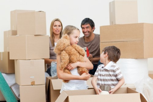 A family of four, including a woman, a man, a young girl holding a teddy bear, and a boy, are inside a residential property surrounded by packed cardboard boxes of various sizes, some stacked on top of each other, indicating a house move. The woman and man are smiling as the girl, standing in front of them, interacts with the boy who is seated in a partially opened box. The boxes and household items suggest preparations for relocation within or from Notting Hill or nearby areas. The background shows part of a clean, well-lit interior space with neutral walls, and the scene captures a moment of engagement during the house removals process, aligned with services offered by Notting Hill Man with Van.