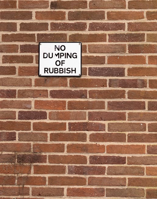 A white rectangular sign with black lettering that reads 'NO DUMPING OF RUBBISH' is mounted on a red brick wall. The bricks are arranged in a traditional horizontal pattern, with some darker bricks sc