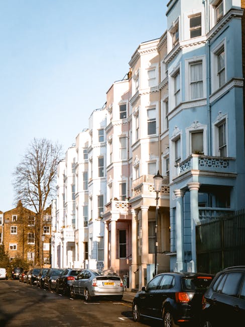 A row of multi-storey residential buildings with ornate white facades and large bay windows, situated along a narrow street in Notting Hill. Several parked cars line the street, including compact and