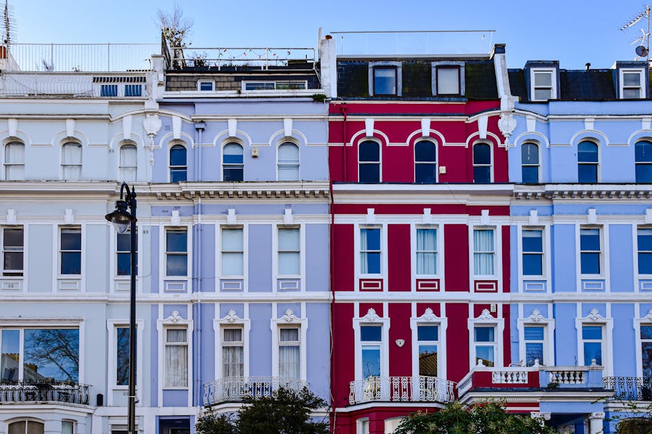 A row of multi-storey residential buildings with ornate white facades and large bay windows, situated along a narrow street in Notting Hill. Several parked cars line the street, including compact and