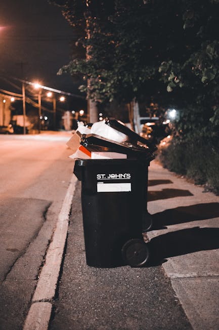 A black wheelie bin labeled 'St. John's' placed upright on a residential pavement at night, with the lid open revealing various waste items including cardboard boxes, plastic packaging, and paper. The