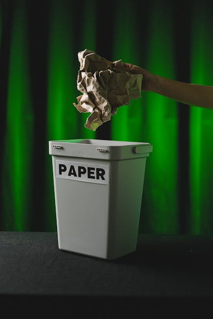A black wheelie bin labeled 'St. John's' placed upright on a residential pavement at night, with the lid open revealing various waste items including cardboard boxes, plastic packaging, and paper. The