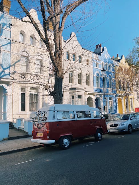 A white moving van parked on a city street alongside a large, multi-storey building with classic architectural features. The van's rear doors are open, revealing a variety of packing materials, includ
